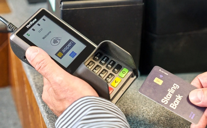  A person holds a bank card near a card payment terminal displaying the Aspen Payments logo, preparing to make a contactless payment at a retail counter 