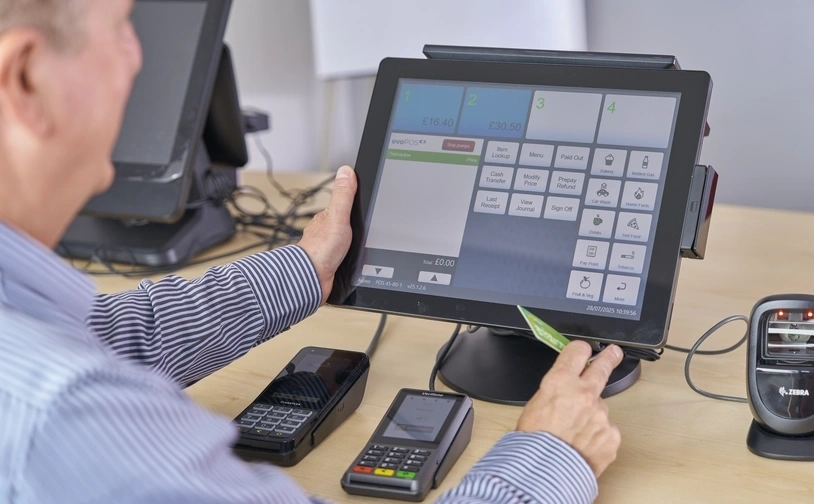 Pete Collett operates the Aspen Payments system dashboard, using a touchscreen point-of-sale terminal with payment devices and a barcode scanner on the desk.