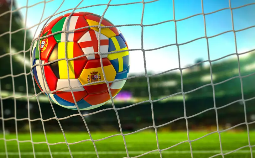 A football decorated with national flags, caught in a goal net against a clear blue sky. Flags include Portugal, Italy, Spain and England