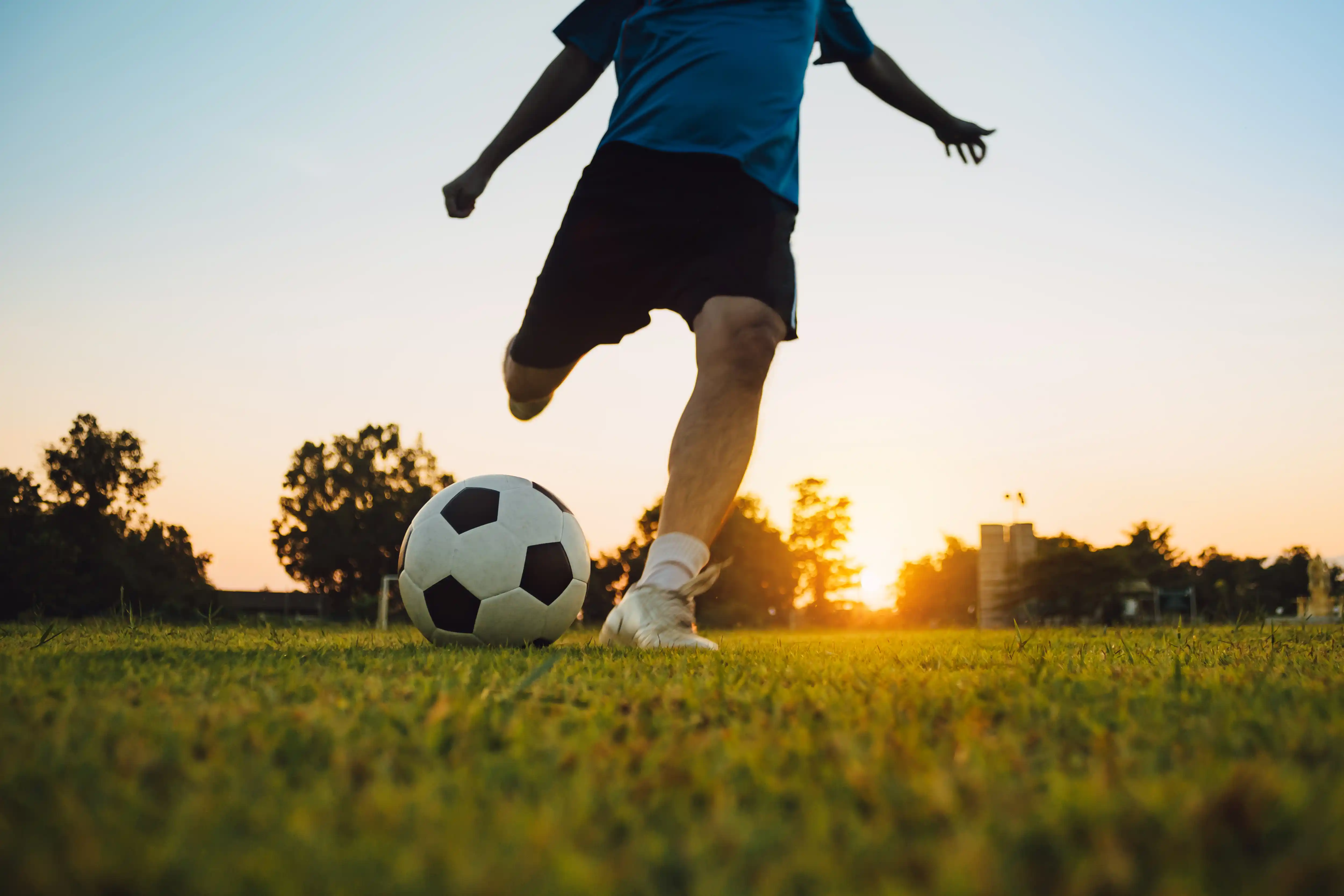 Person kicking a soccer ball on a grassy field during sunset with trees and buildings silhouetted in the background